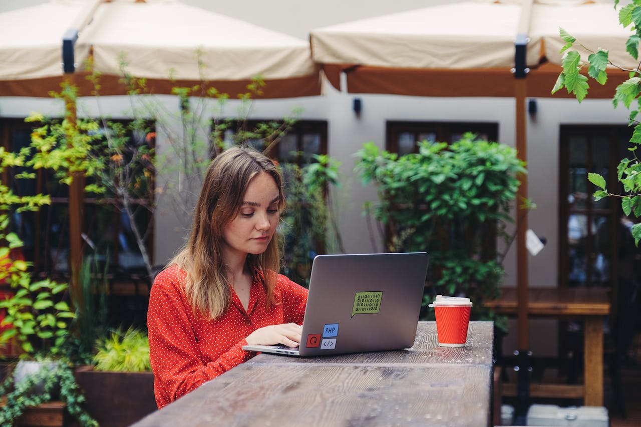 Young woman working on a laptop in an outdoor cafe setting, enjoying a cup of coffee.